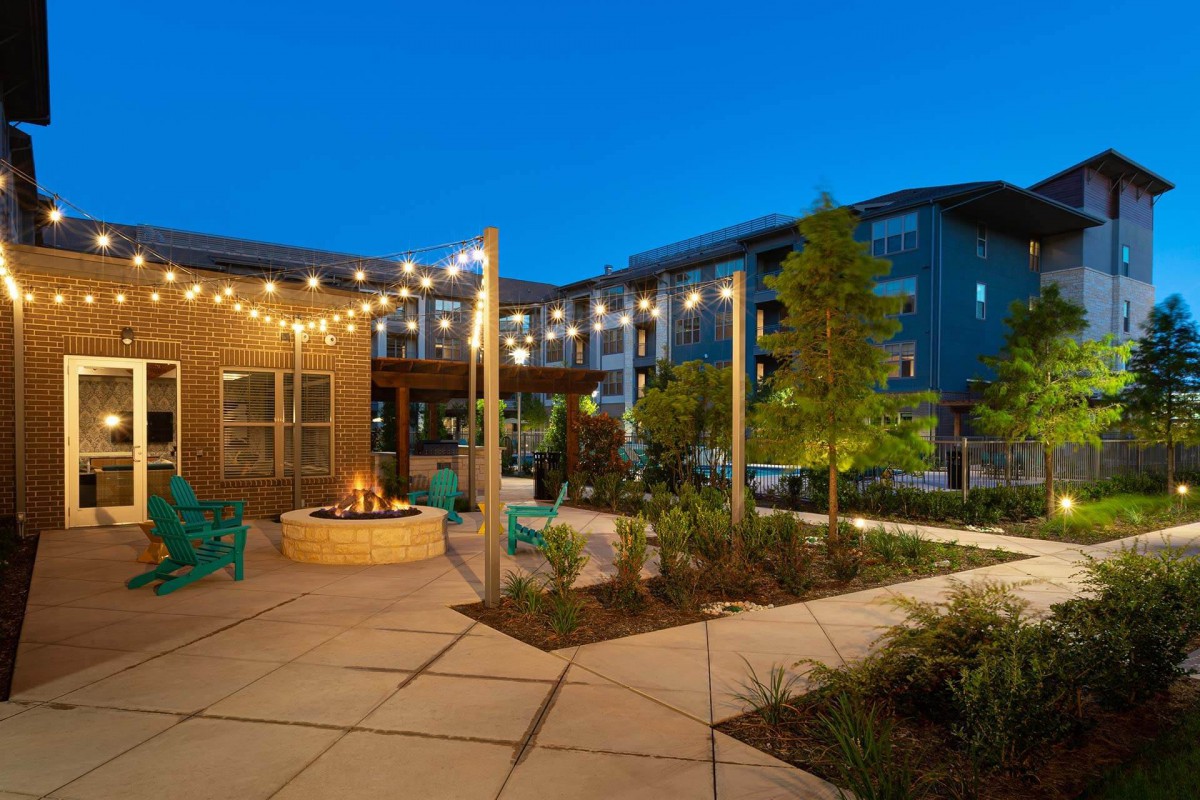A modern apartment complex with a beautifully landscaped outdoor area, featuring string lights and a fire pit, captured during the evening.