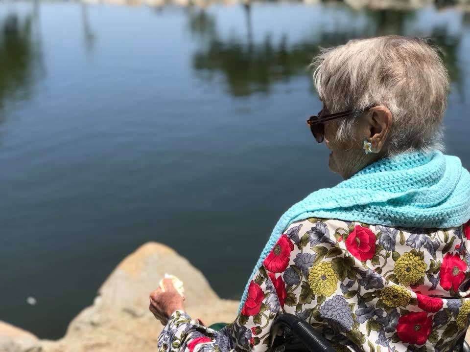An elderly woman wearing sunglasses and a floral shawl, sitting by a lake enjoying the outdoors.