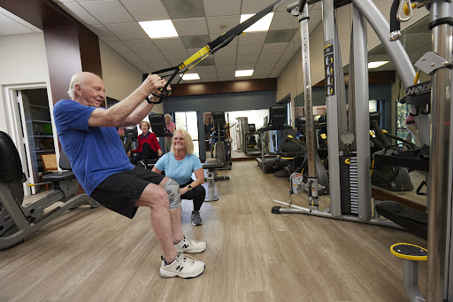 An older adult engages in a TRX workout, supported by a trainer in a vibrant gym environment.