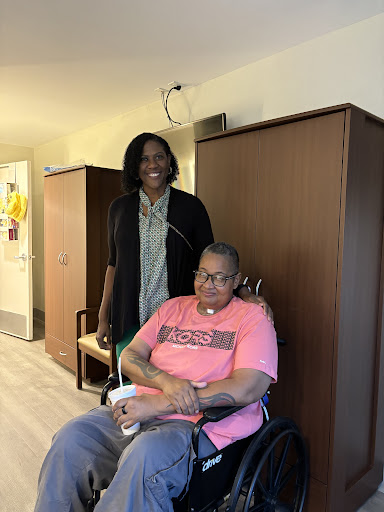 A caregiver stands beside a person in a wheelchair, both smiling in a comfortable indoor setting.