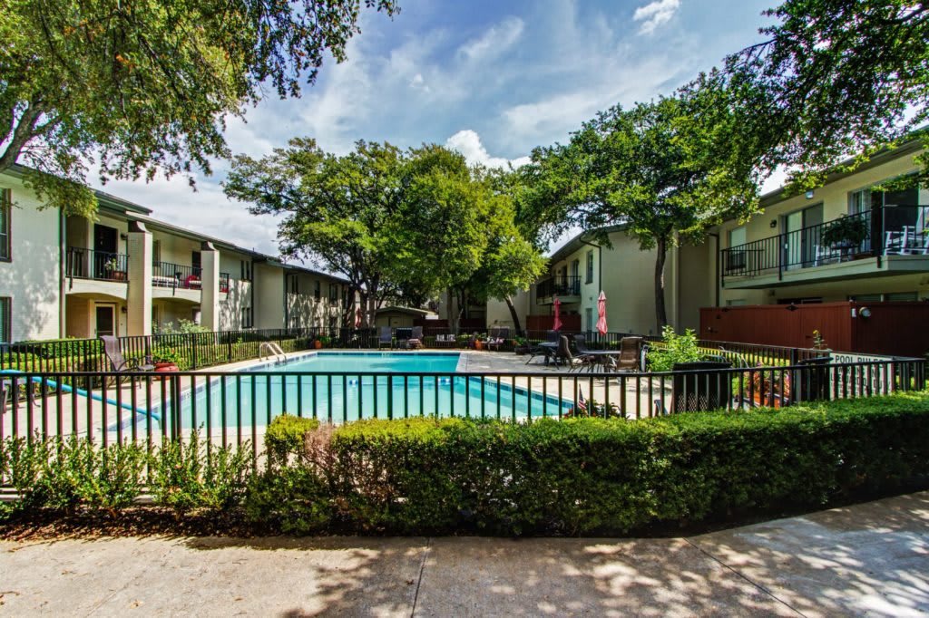 A serene view of an apartment complex featuring a fenced swimming pool surrounded by lush greenery on a sunny day.
