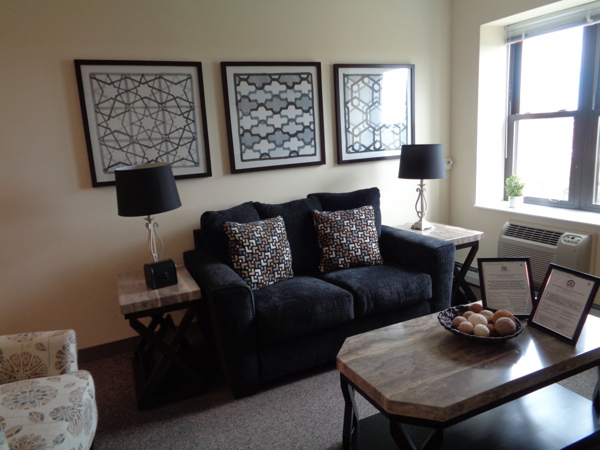 A cozy living room with a dark sofa, decorative cushions, and framed artwork above it. The space is complemented by side tables with lamps and a central coffee table.