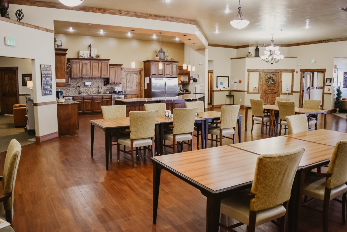 A spacious and well-lit dining area with a kitchen, featuring wooden tables and chairs on a polished wooden floor.