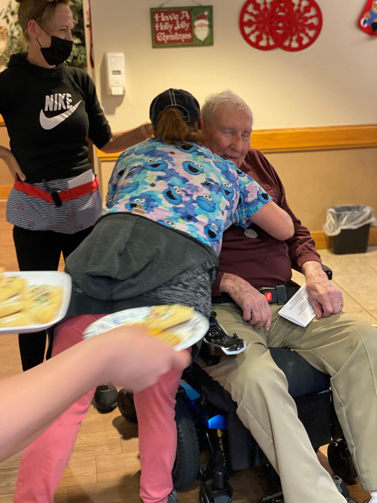 An elderly man in a wheelchair receives a hug from a caregiver while another person holds plates of food.