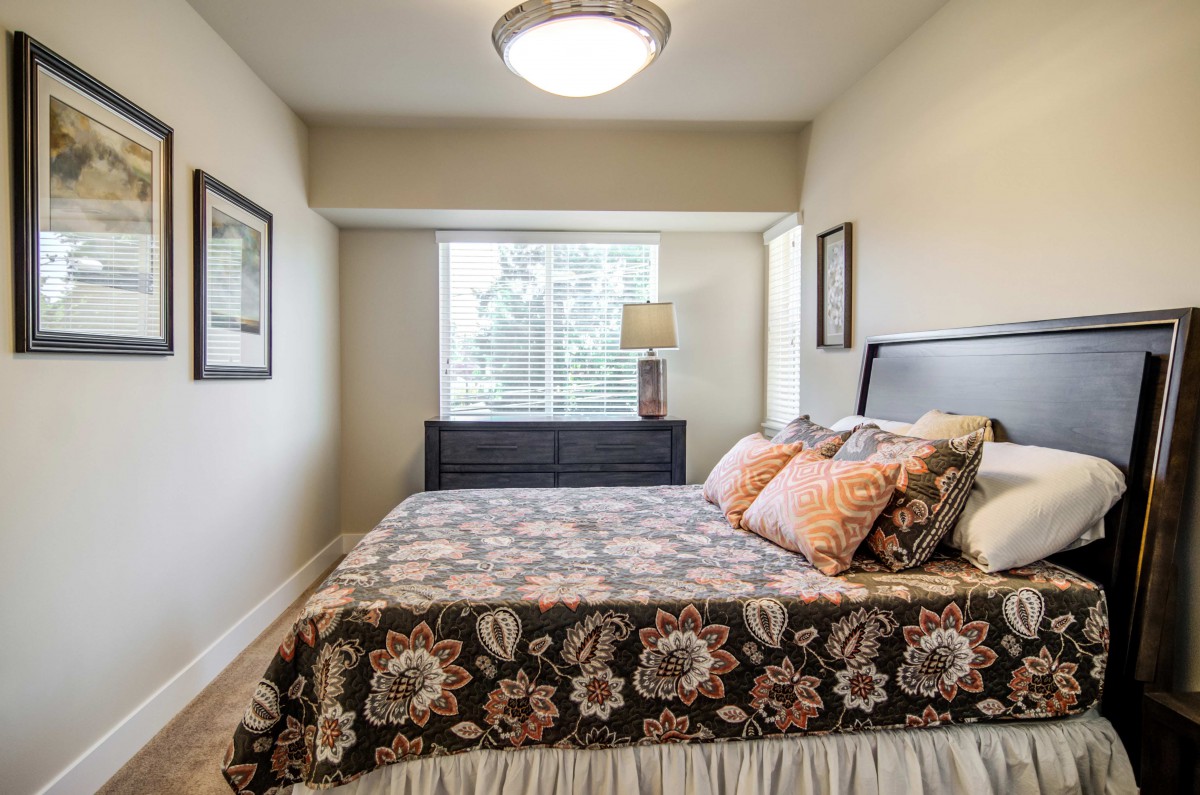 A cozy bedroom featuring a bed with floral bedding, a lamp on the dresser, and natural light streaming through the window.