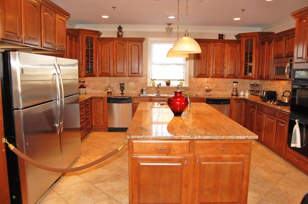 A spacious kitchen featuring wooden cabinets, a central island with a red vase, stainless steel appliances, and pendant lighting.