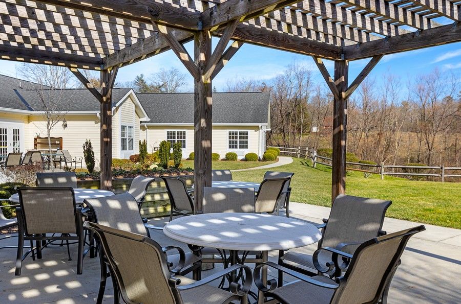 A sunlit patio area with tables and chairs under a pergola, overlooking a landscaped garden and building in the background.