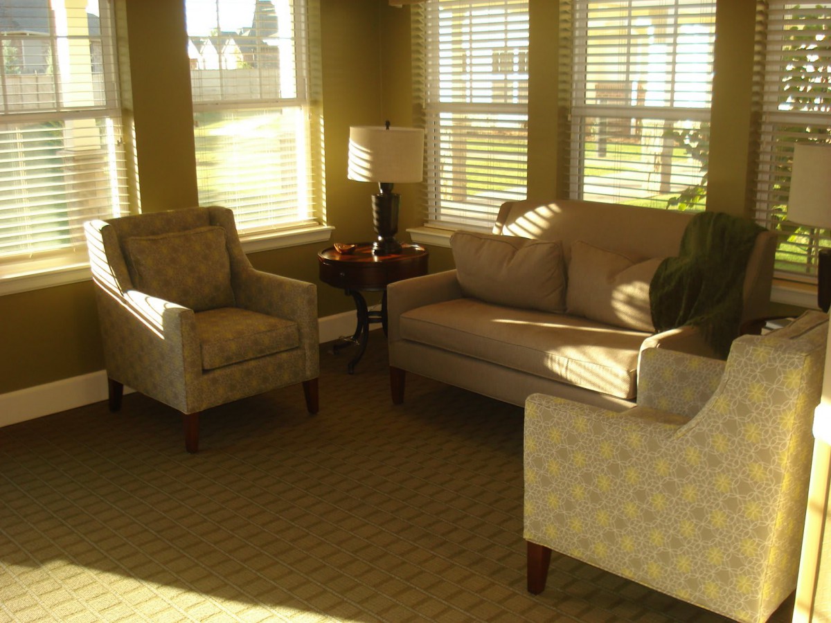 A cozy living room with natural light streaming through the windows, featuring a sofa, armchairs, and a lamp.