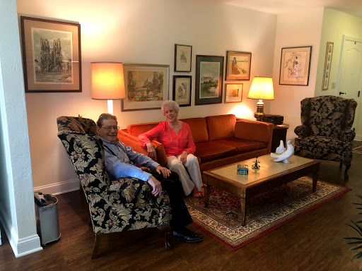 A cozy living room featuring a couple seated together on a patterned armchair and sofa, surrounded by art and warm lighting.