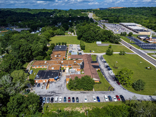 Aerial view of a building complex surrounded by greenery and a parking lot, with a roadway visible in the distance.