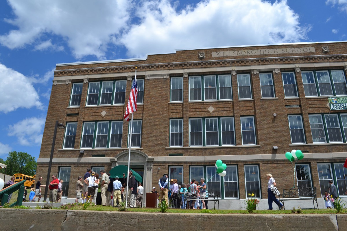 A group of people gathered outside a large brick building on a sunny day, with an American flag and green balloons visible.