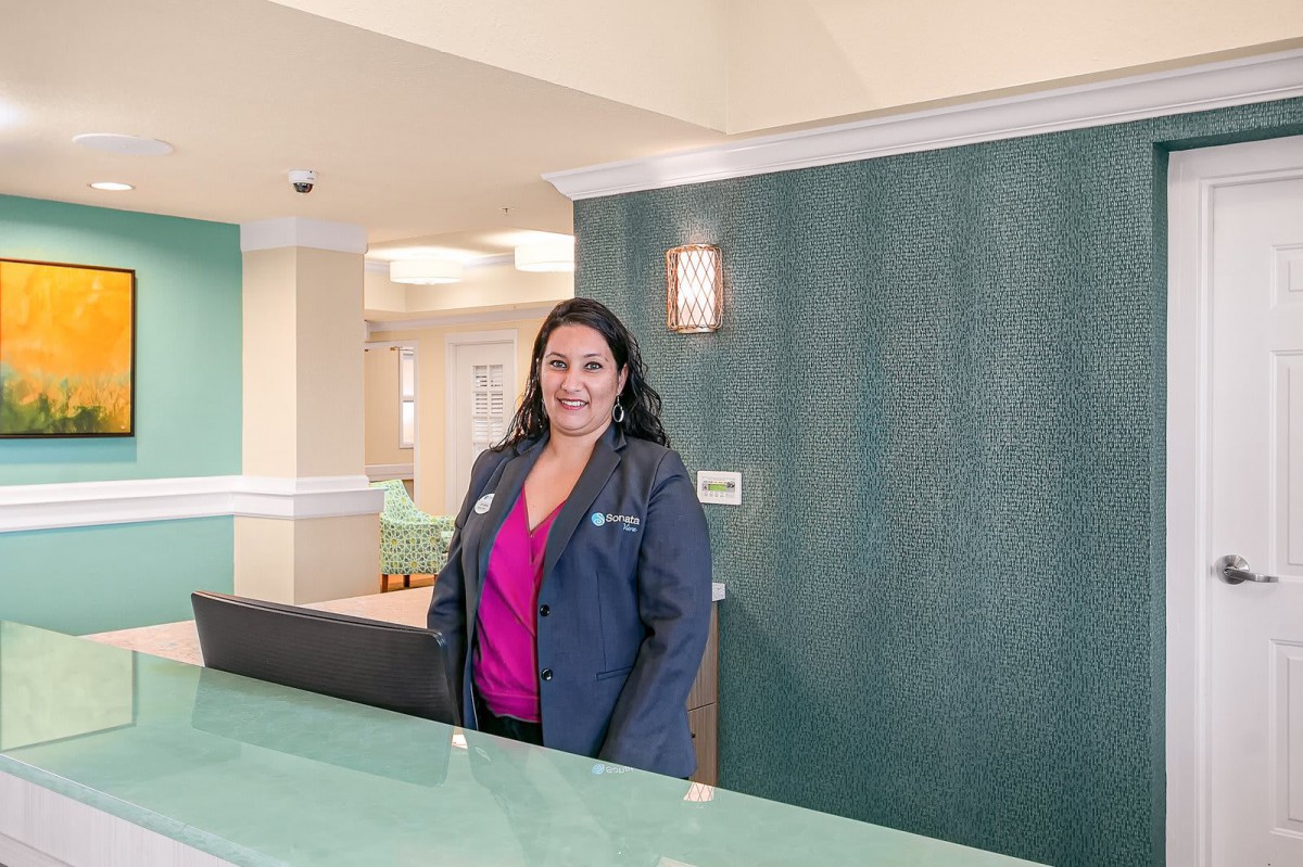 A receptionist standing behind a desk in an office environment, smiling at the camera.