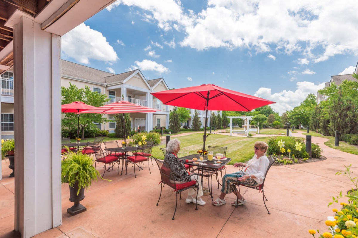 Seniors enjoying a sunny day on the patio with red umbrellas and garden views.