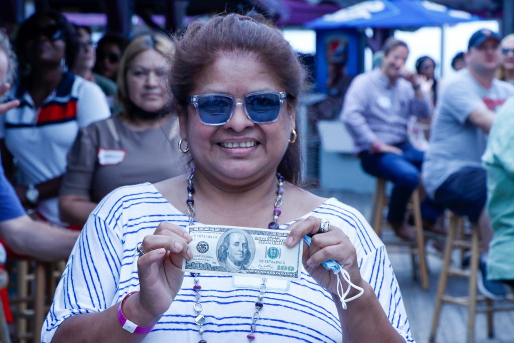 A woman wearing sunglasses smiles while holding a dollar bill at an outdoor event.