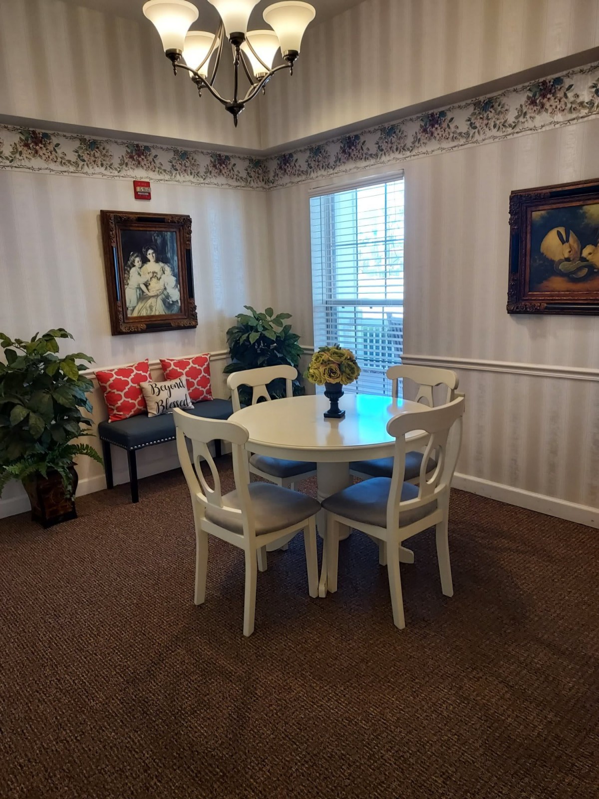 A cozy dining corner featuring a round table with four chairs, adorned with plants and paintings, next to a large window.