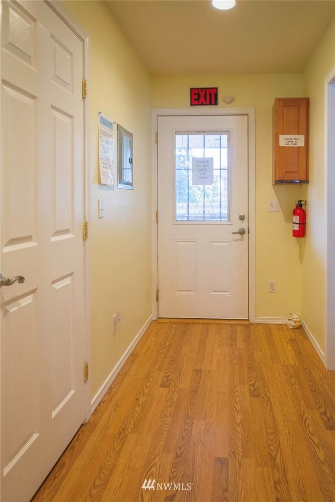 A hallway with wooden floors leading to a door with an exit sign above it and a fire extinguisher to the side.