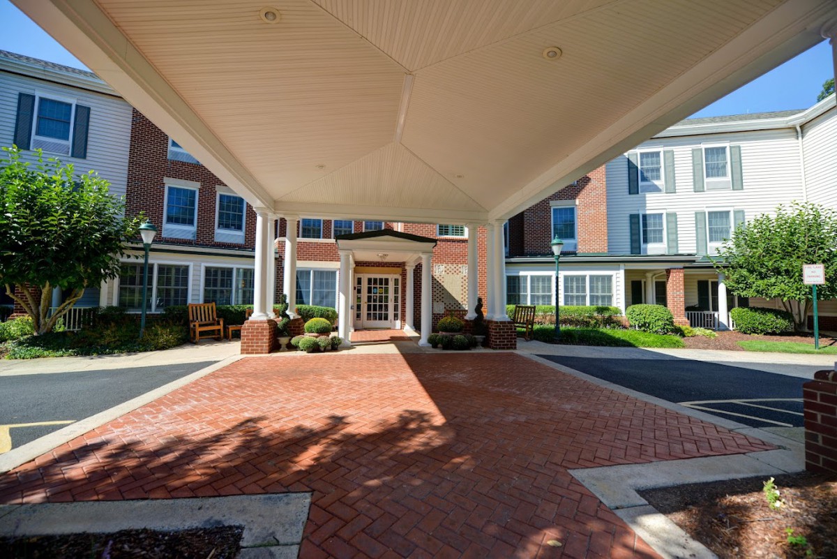 The front entrance of a building with a covered driveway and brick pathway, surrounded by well-maintained landscaping.