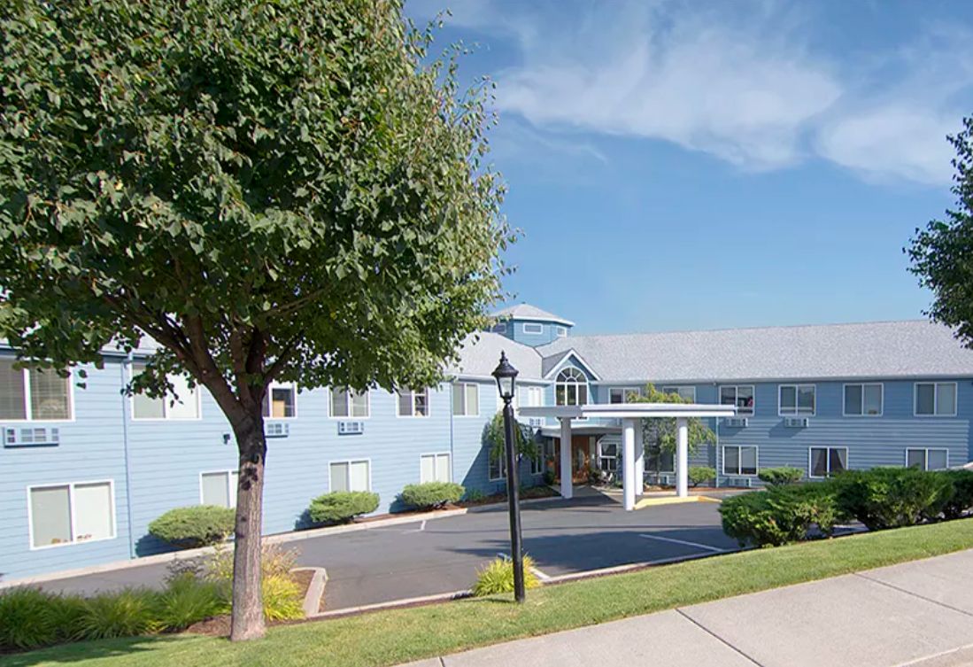 A two-story building with a circular driveway, surrounded by greenery and set against a clear blue sky.