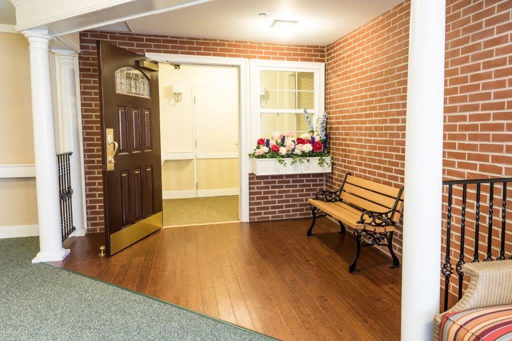 An interior space with a wooden door, adjacent brick wall, window with flower box, and a decorative bench.