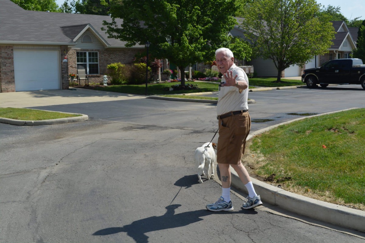 An elderly man walking his dog in a residential neighborhood on a sunny day.