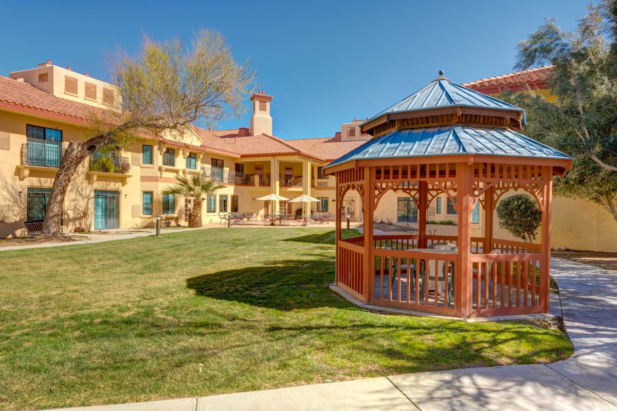 A sunny courtyard featuring a wooden gazebo and a surrounding building.