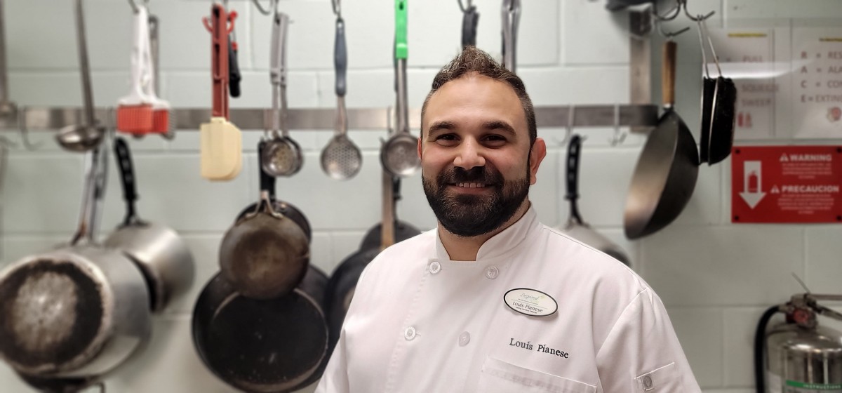 A chef smiling in a kitchen with cooking utensils hanging in the background.