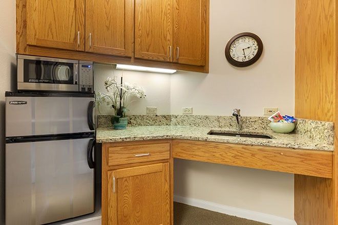 A small kitchen area with wooden cabinets, a granite countertop, and a sink. A clock is mounted on the wall above, and an orchid plant decorates the counter next to a mini refrigerator.