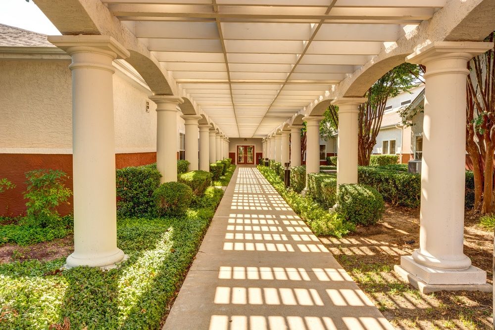 A sunlit corridor with columns casting shadows on a covered walkway surrounded by greenery.