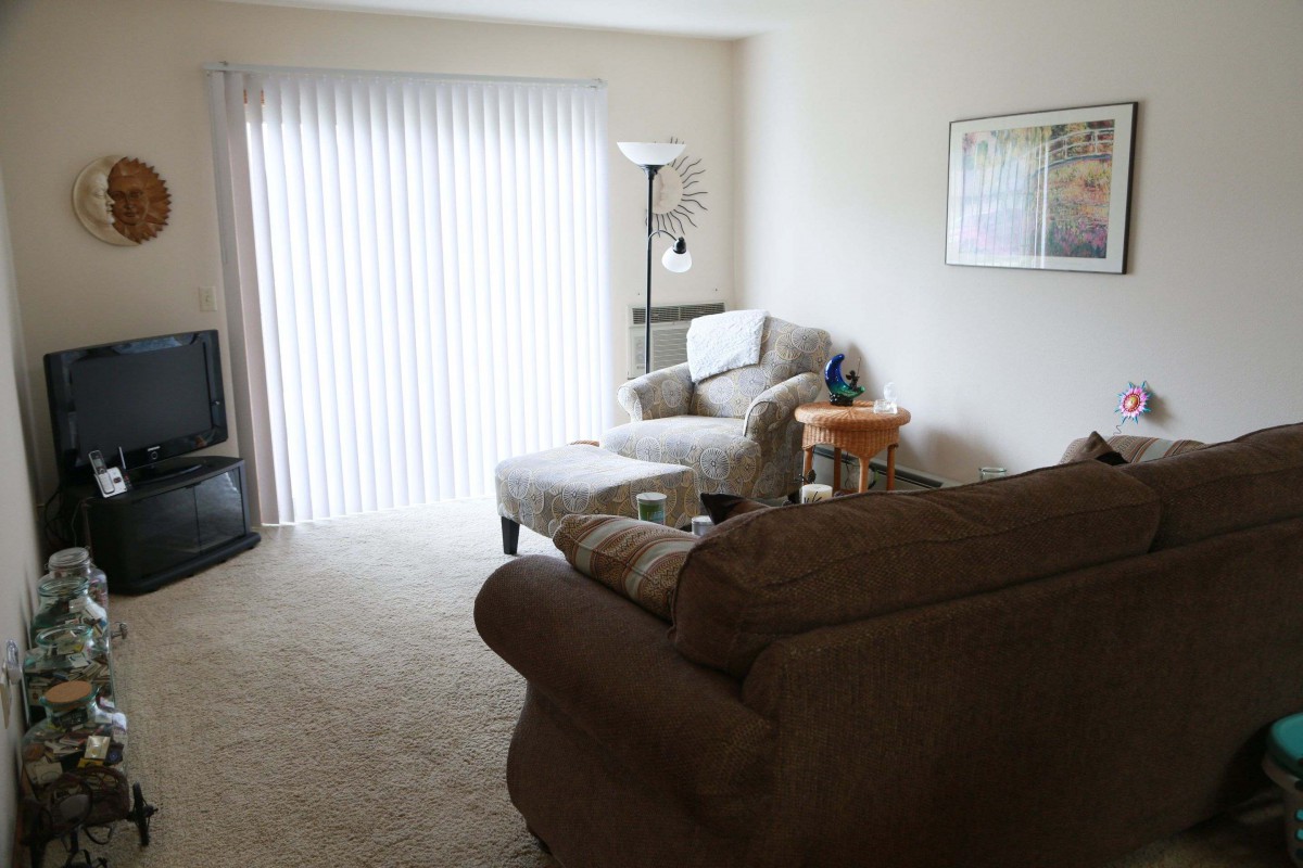 A cozy living room with soft lighting, featuring a sofa and chair arranged around a small table, with window blinds letting in natural light.