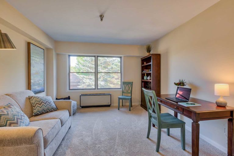 A well-lit home office with a desk, chair, sofa, and bookshelf.