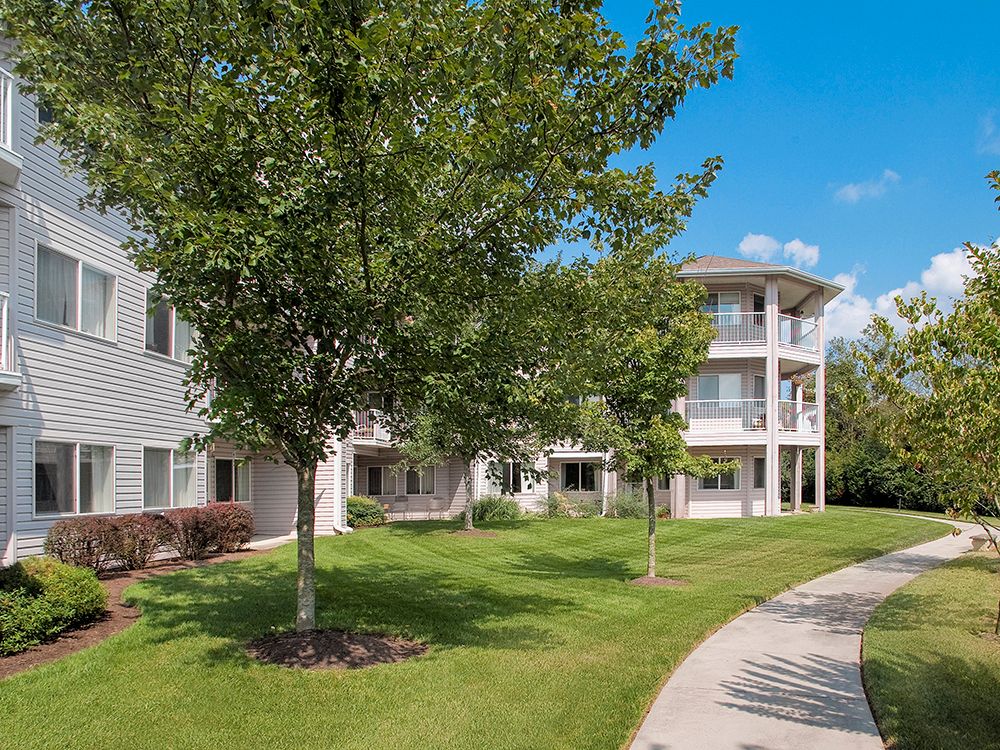 A residential apartment complex with landscaped lawns, trees, and a pathway on a sunny day.