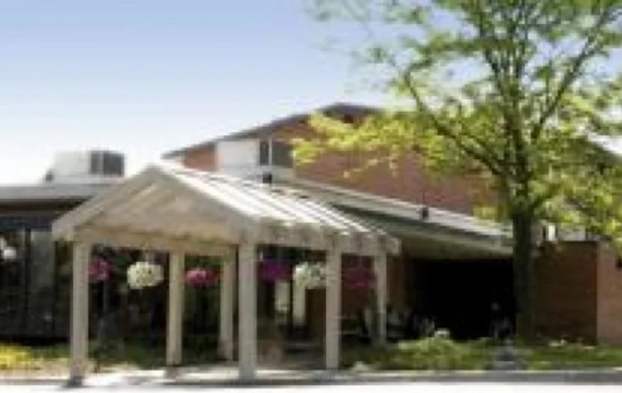A building entrance with a glass canopy, surrounded by trees and flowers.