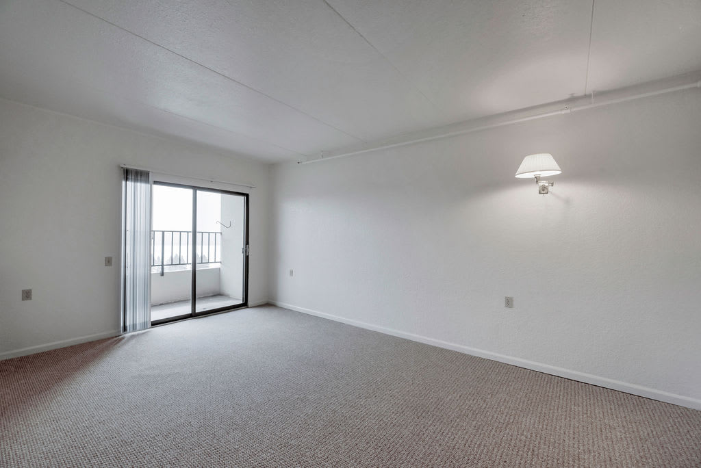 An empty room with white walls and carpeted flooring, featuring a sliding door that leads to a balcony.