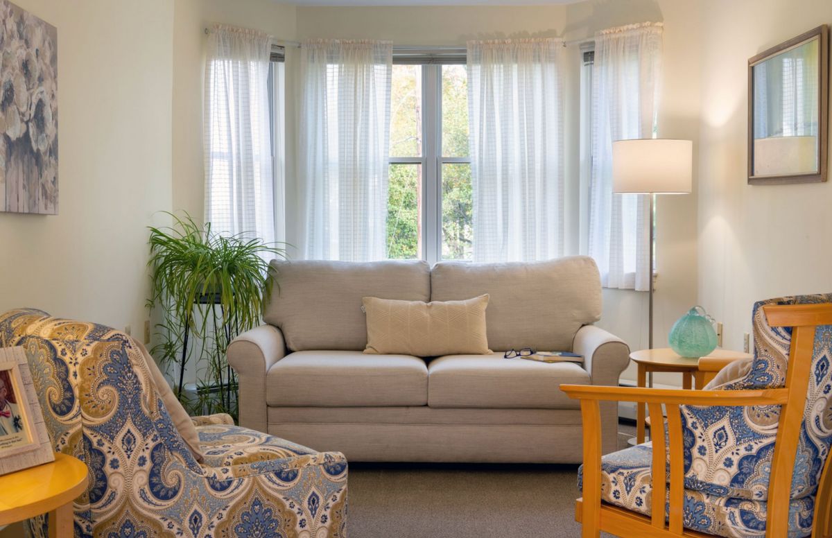 A cozy living room with a sofa, armchair, and floor lamp in front of a window with sheer curtains.