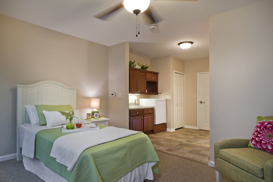 A cozy bedroom with a bed, side table, lamp, and a kitchenette area.