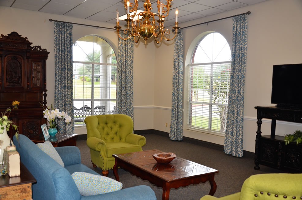 A cozy living room with a chandelier, green armchairs, a blue sofa, and large arched windows with curtains.
