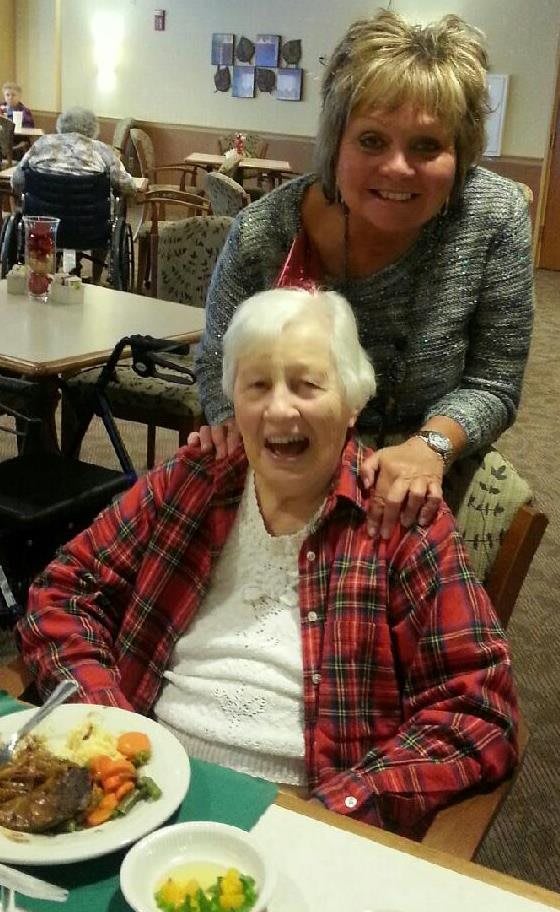 An elderly woman in a plaid shirt is smiling joyfully with another person standing behind her, suggesting a celebratory occasion indoors.