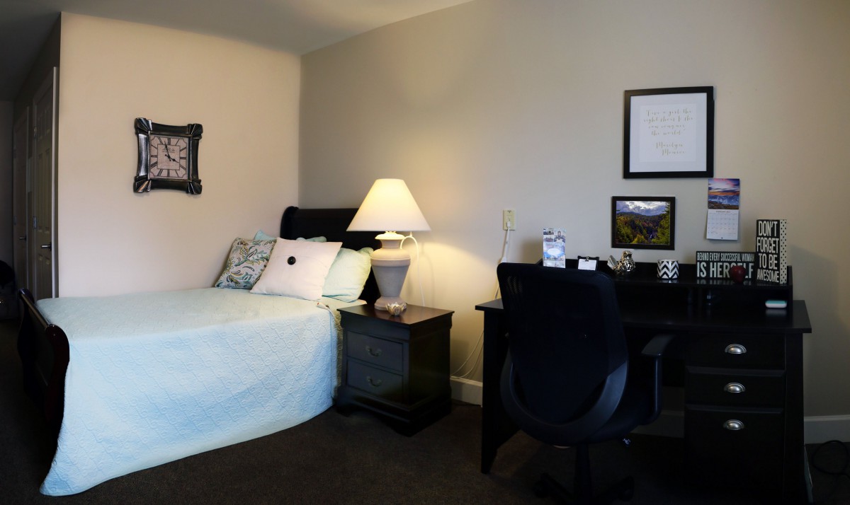 A cozy bedroom with a bed, nightstand, lamp, and a desk with a chair.