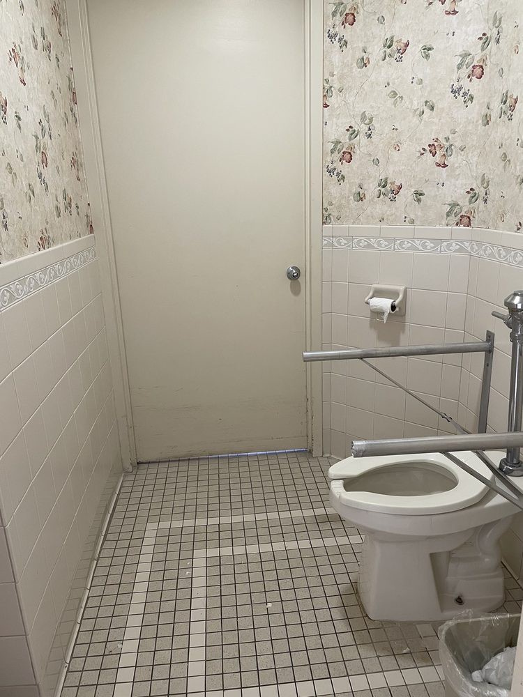 A small bathroom with a tiled floor, floral wallpaper, and a toilet equipped with grab bars.