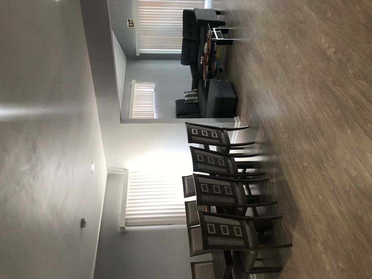 A spacious living room with a dining area, featuring a set of chairs and a table, complemented by large windows and hardwood flooring.