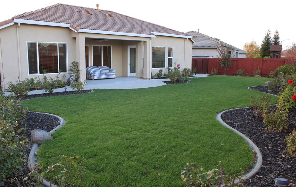 A backyard with a green lawn, patio area, and surrounding garden landscaping.