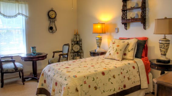 A cozy bedroom with a single bed dressed in floral bedding, illuminated by a warm lamp alongside decorative vintage clocks.