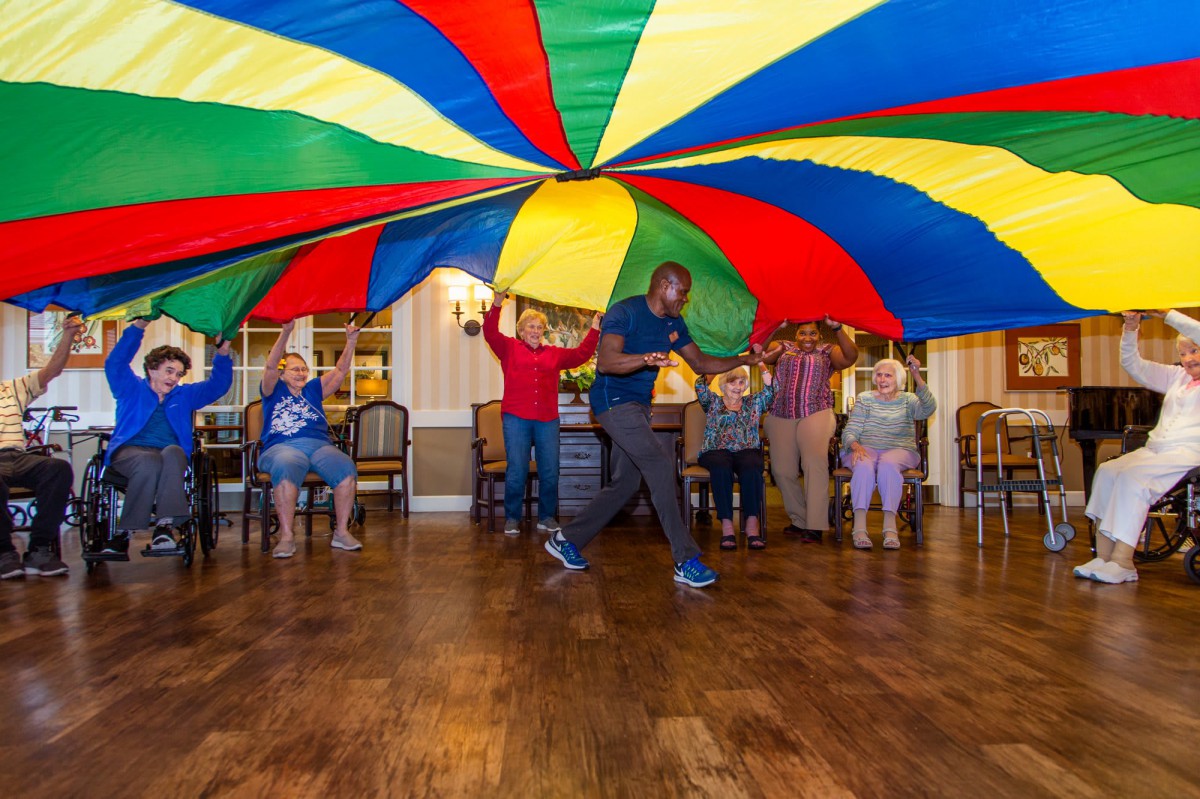 A group of people participating in a fun indoor parachute game, lifting a vibrant and colorful parachute together.