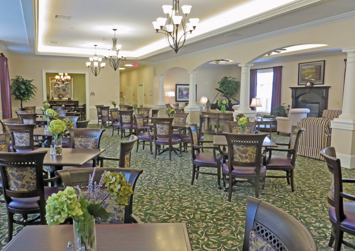 A spacious and elegantly decorated dining room with multiple tables and chairs, chandeliers, and greenery.
