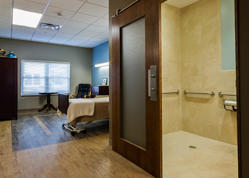 A modern patient room featuring a shower area, a bed, and an office chair by the window.