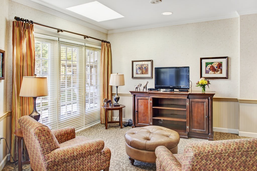 A cozy living room with armchairs, a television on a wooden stand, and large windows with blinds.