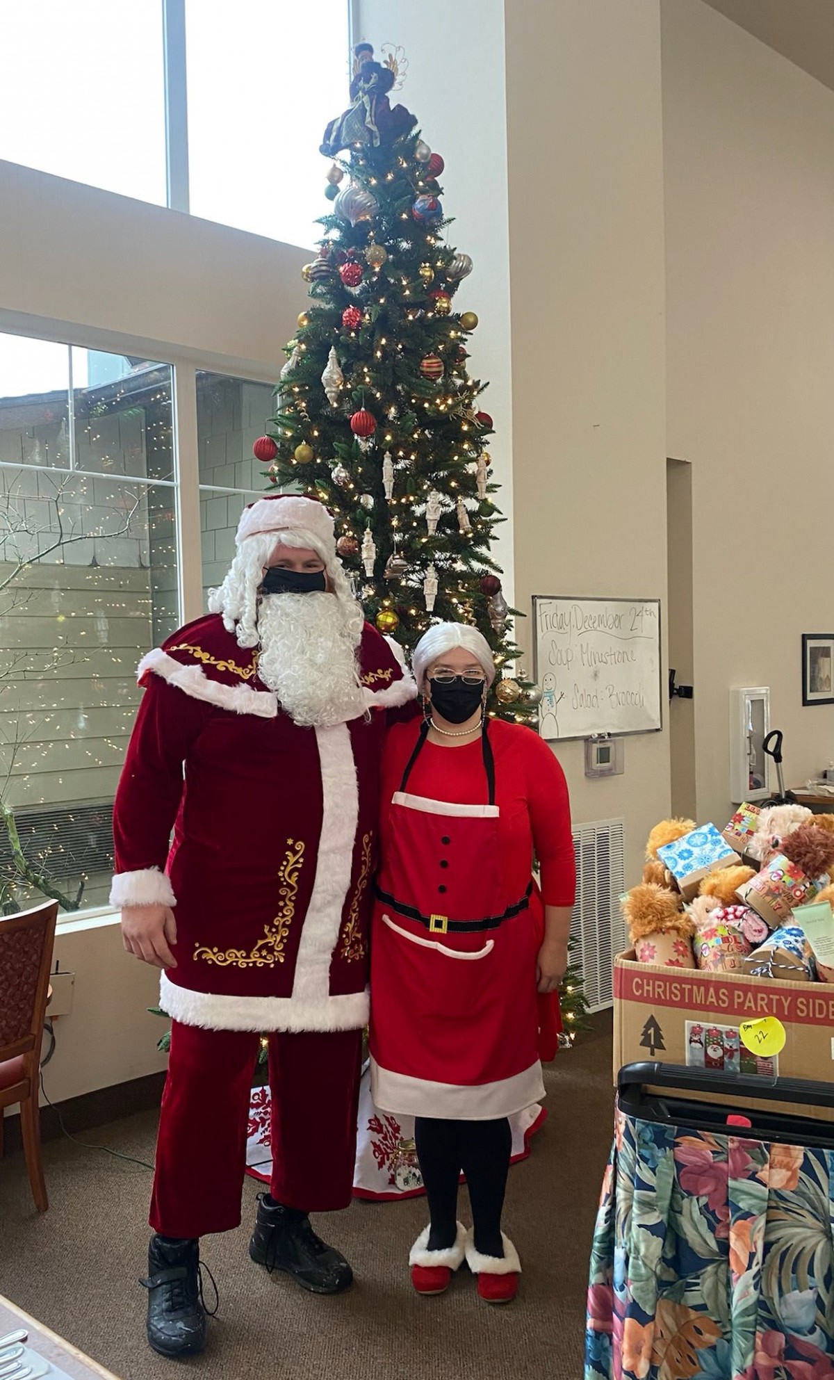 Two people dressed in Santa costumes pose beside a decorated Christmas tree indoors.