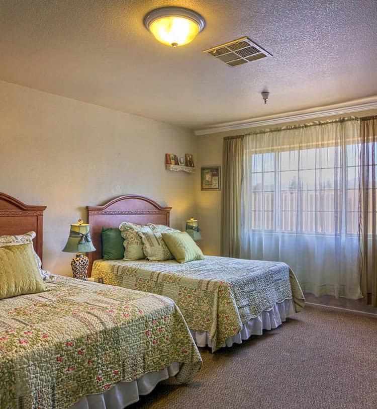 A cozy bedroom featuring two twin beds with matching bedspreads, soft lighting, and natural light streaming through the curtains.