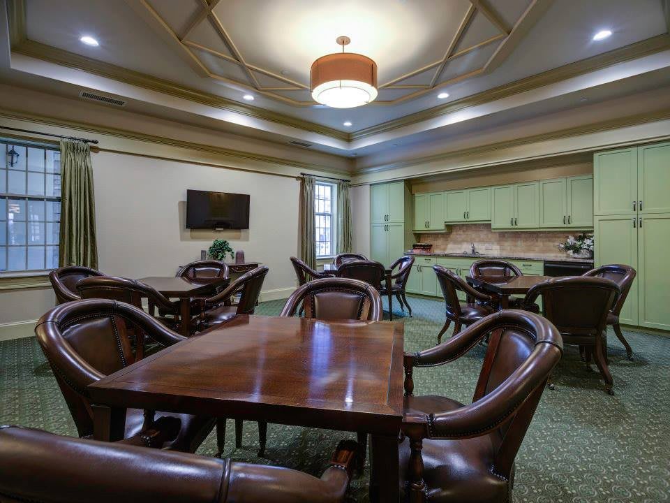 A well-lit dining area featuring several tables and chairs, with a wall-mounted television and a kitchenette in the background.