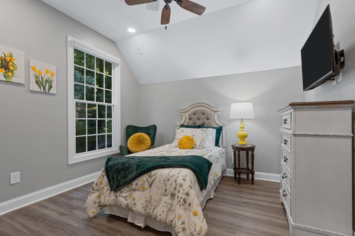 A cozy and neatly arranged bedroom with a single bed, adorned with decorative pillows and a warm blanket. A lamp on the nightstand adds a soft glow, while a window provides natural light.